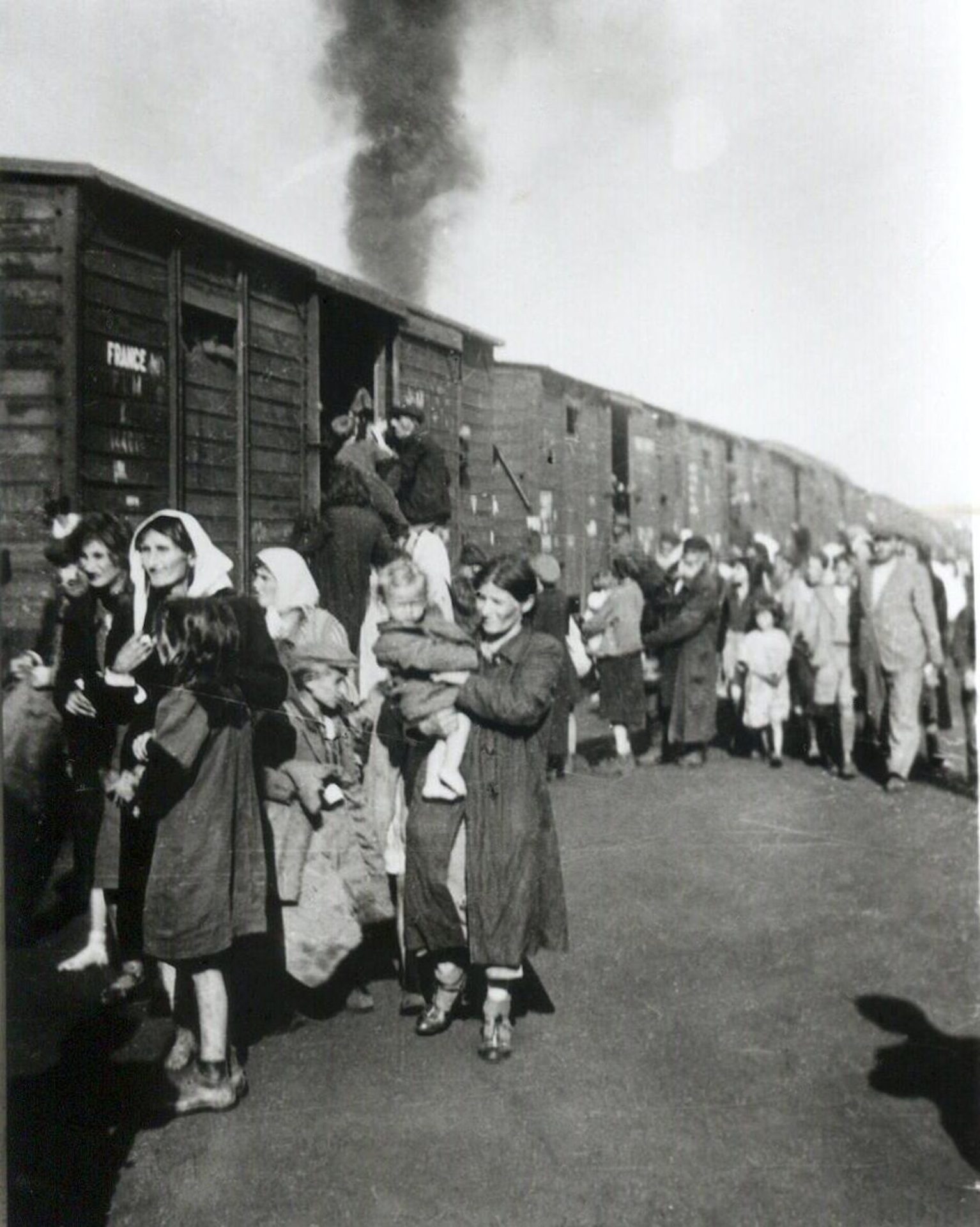 Una foto en blanco y negro muestra a mujeres y niños con abrigos caminando junto a vagones de ganado.