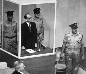 29/5/1962-Jerusalén, Israel- Adolf Eichmann, acusado de asesino en masa nazi, se para en su jaula de vidrio a prueba de balas para escuchar a la Corte Suprema de Israel rechazar por unanimidad una apelación contra su sentencia de muerte. Con él hay dos guardias armados. El veredicto significó que solo el presidente israelí Itzhak Ben-Zvi podía salvar a Eichmann, de 56 años, acusado del asesinato de seis millones de judíos, de la horca. En primer plano está el abogado defensor Robert Servatius.